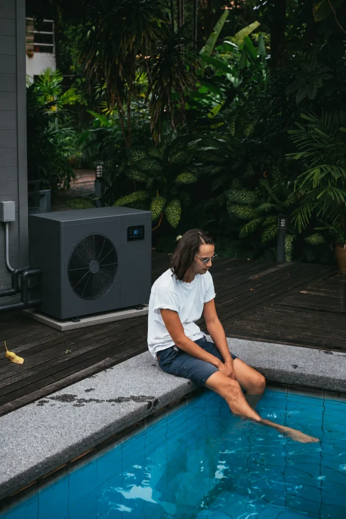 Modern pool heat pump installed beside a backyard swimming pool, with a homeowner relaxing by the water in a lush outdoor setting